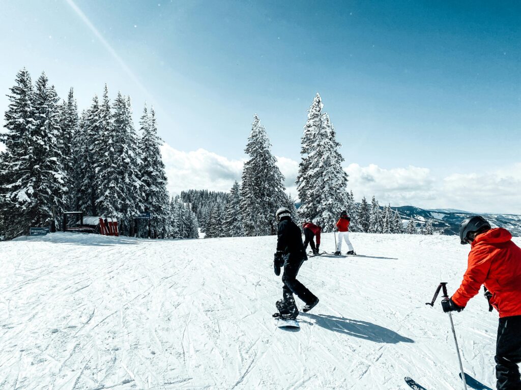 A group of snowboarders and skiers enjoying a sunny winter day on a snowy slope surrounded by trees.
